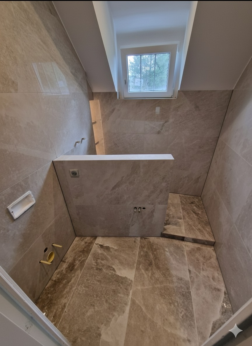 Wykończona łazienka w beżowych płytkach imitujących marmur, z oknem w skosie. Widoczne przygotowane instalacje wodne i elektryczne, oraz fragment niedokończonej podłogi.