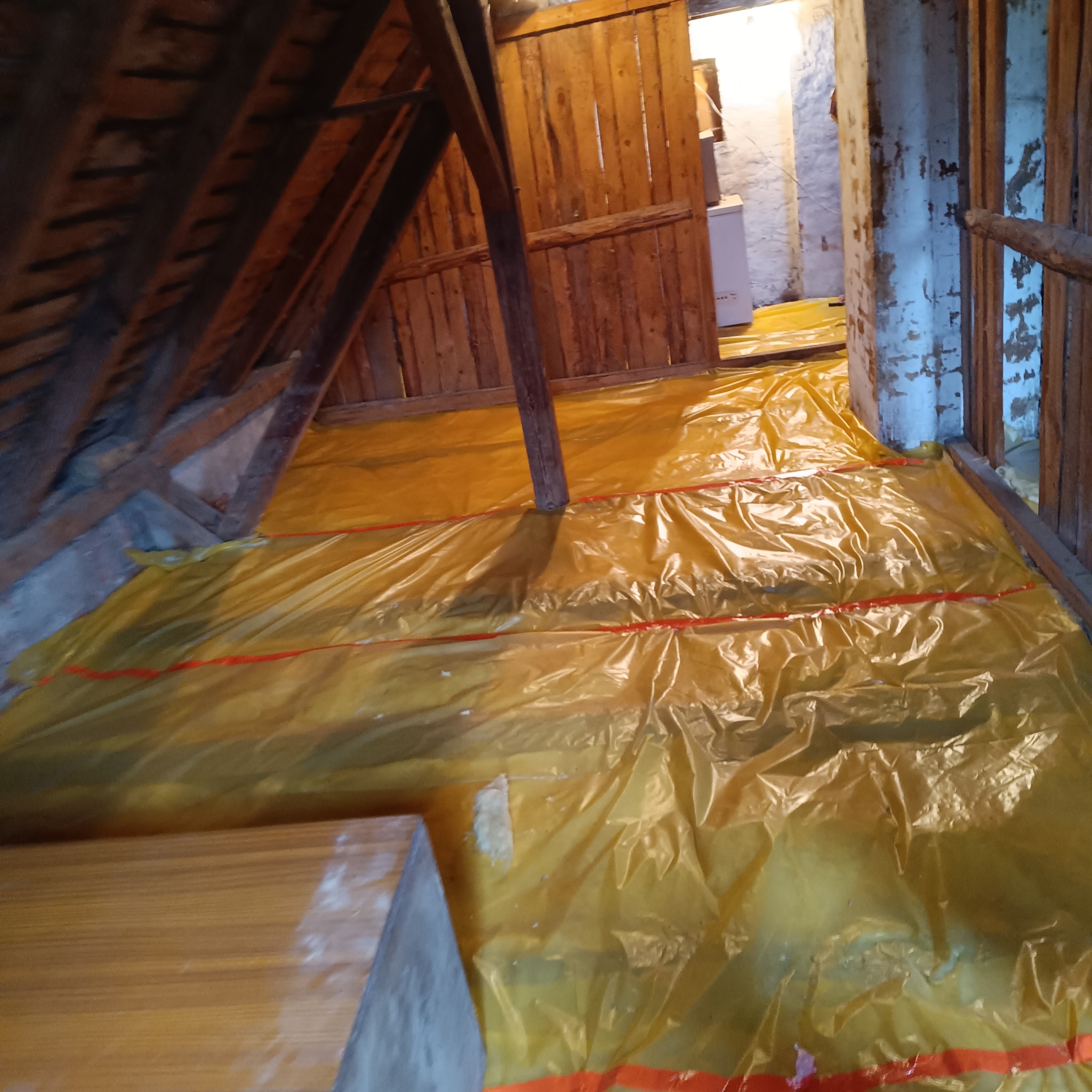 Poddasze zabezpieczone żółtą folią budowlaną przed rozpoczęciem prac izolacyjnych, widoczne drewniane elementy konstrukcyjne dachu i fragment ściany z widocznymi śladami starej farby.