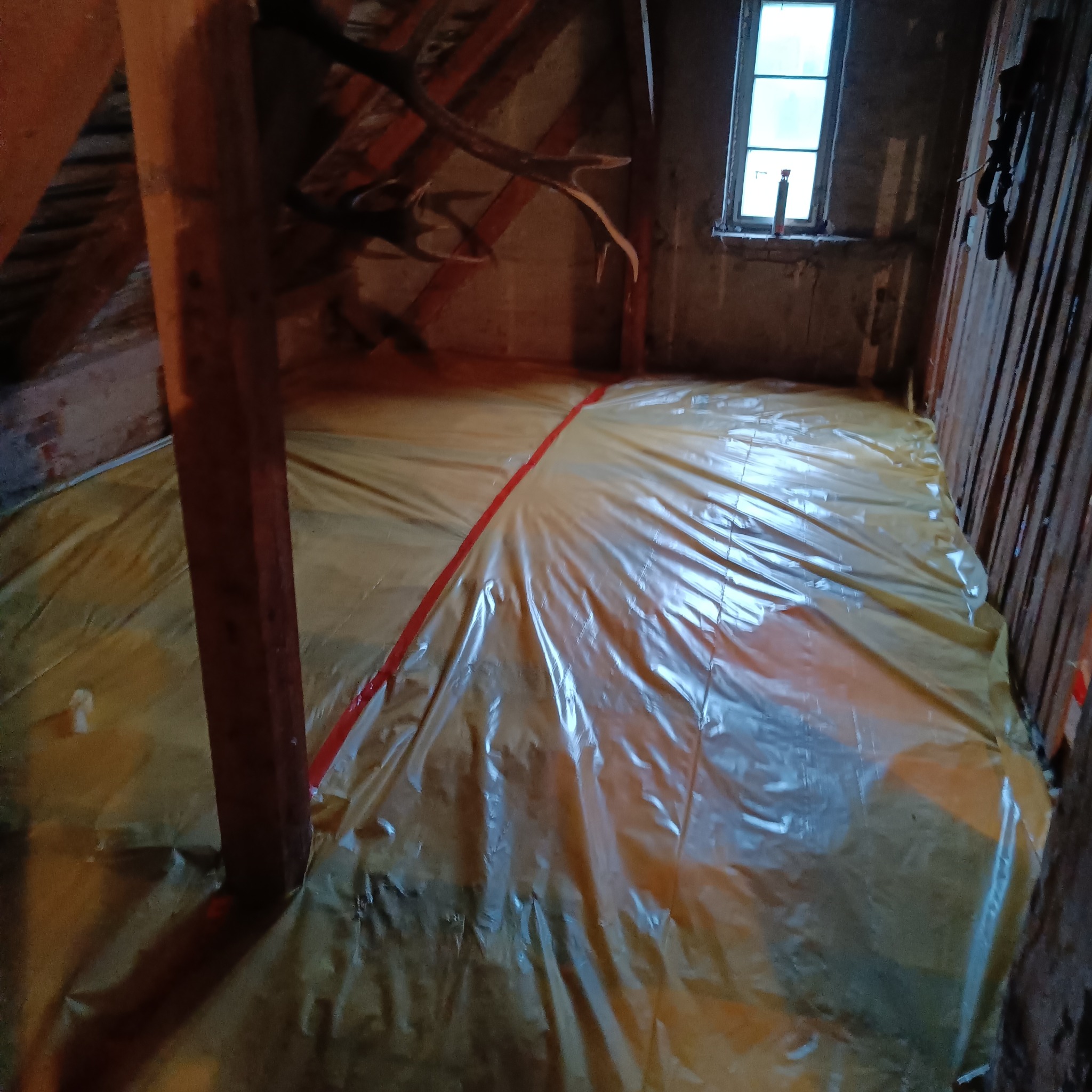 Przygotowanie poddasza do izolacji: żółta folia ochronna rozłożona na podłodze, drewniane elementy konstrukcyjne więźby dachowej, okno w tle.