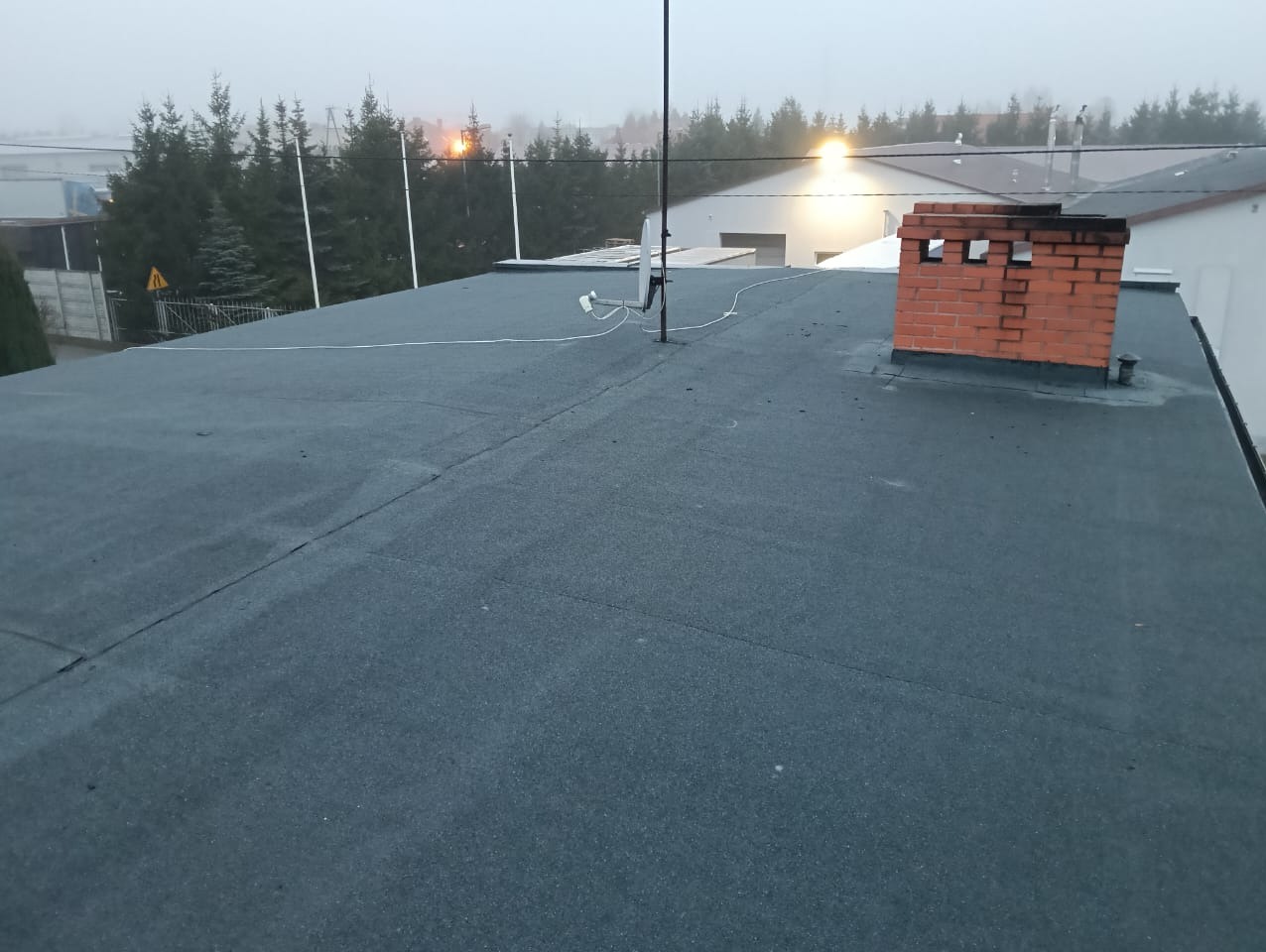 Płaski dach pokryty czarną papą termozgrzewalną z widocznym kominem z cegły, anteną i elementami instalacji odgromowej na tle zamglonego krajobrazu.