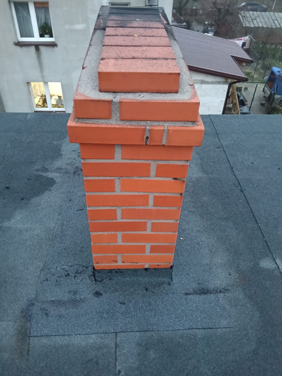 Murowany komin z czerwonej cegły wystający z pokrytego papą dachu, widoczny fragment budynku mieszkalnego w tle.