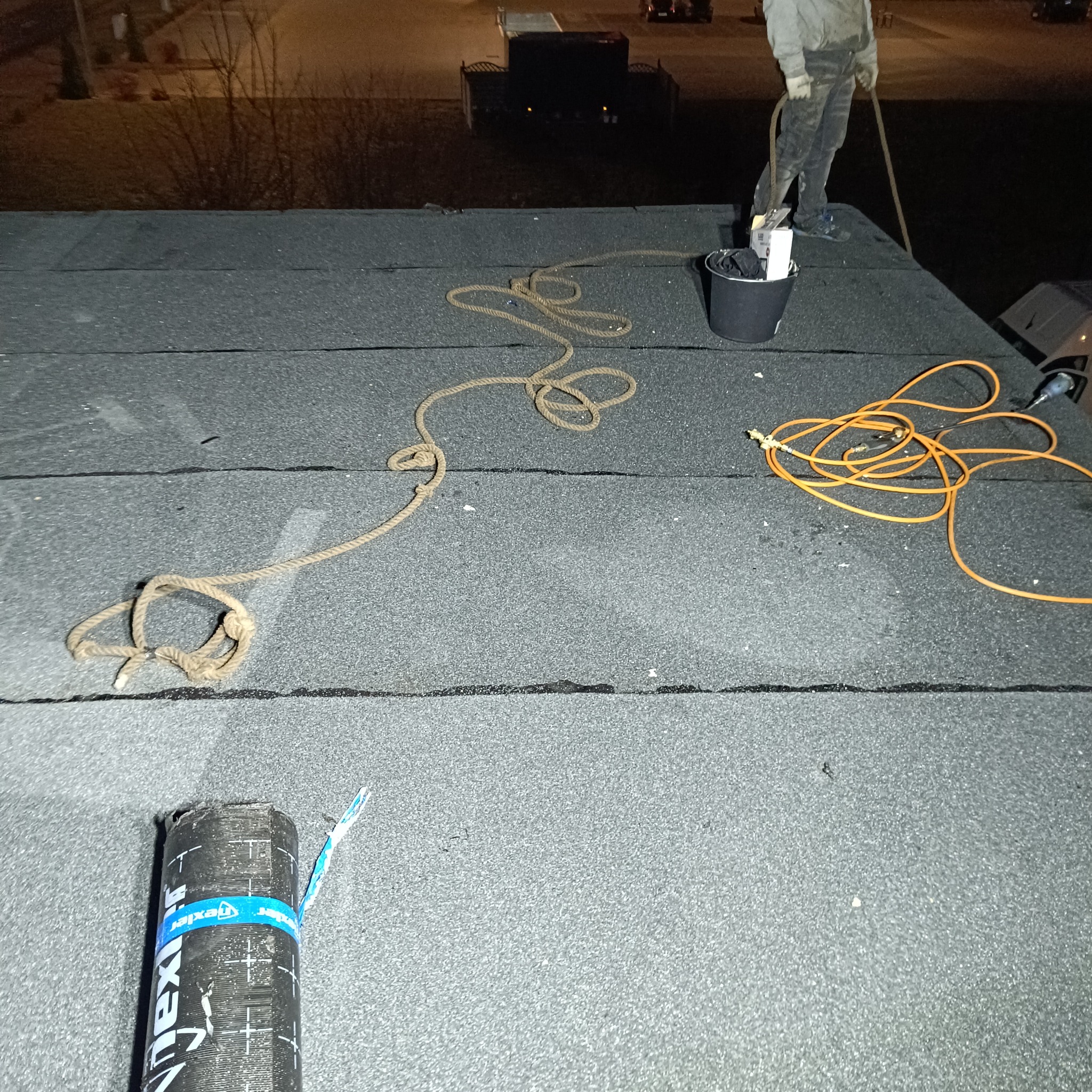 Prace na dachu pokrytym papą termozgrzewalną w nocy, widoczny pracownik z wiadrem, zwój papy Dexter i lina zabezpieczająca.