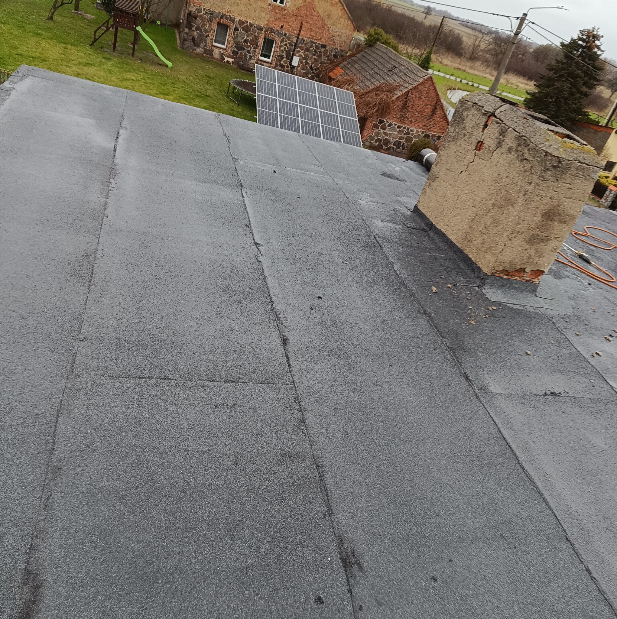 Widok z góry na płaski dach pokryty papą termozgrzewalną w Milanówku, z kominem w tle i instalacją fotowoltaiczną na trawniku.