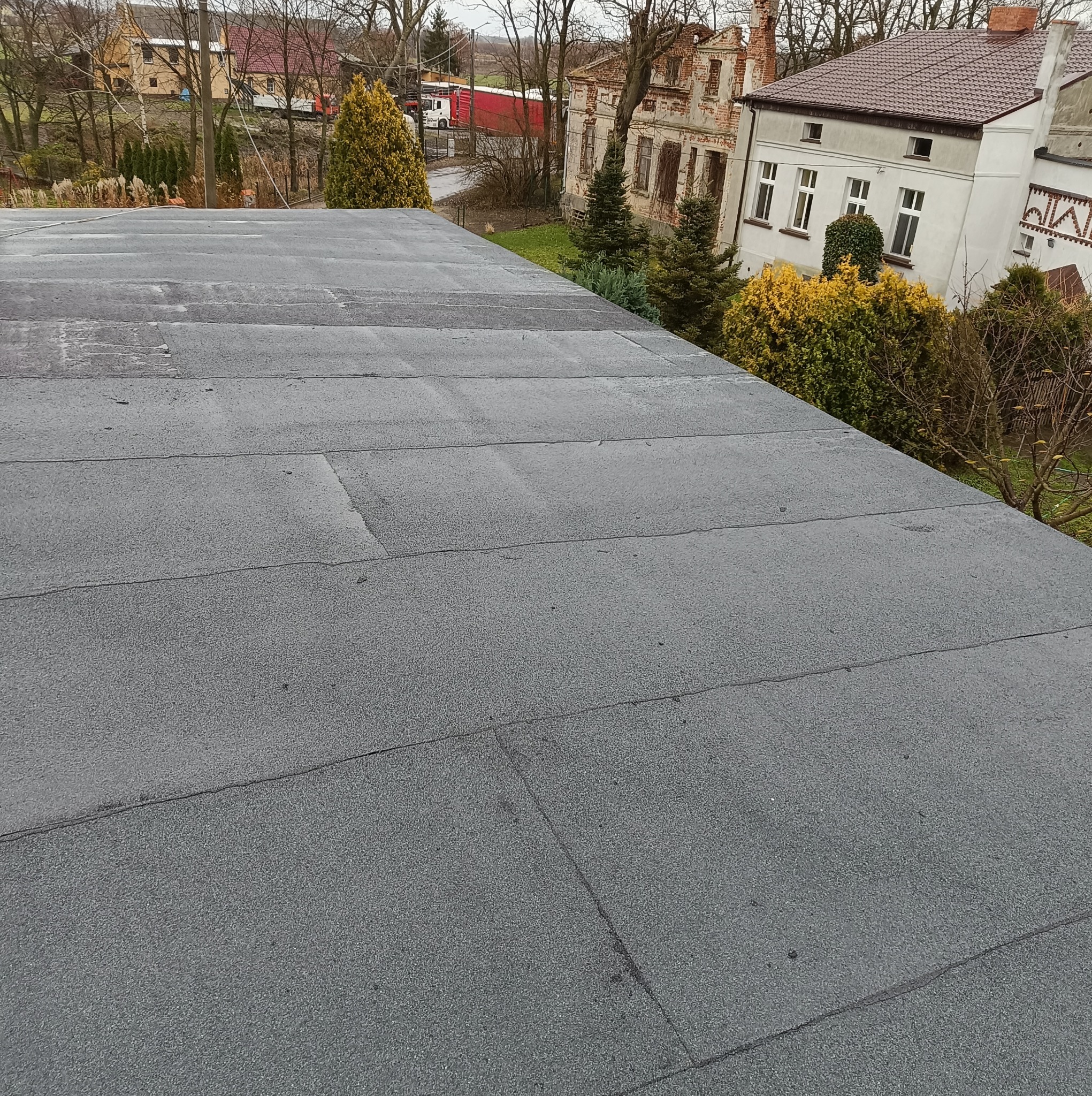 Szary, płaski dach pokryty papą termozgrzewalną z widocznymi łączeniami, w tle budynki mieszkalne i drzewa.