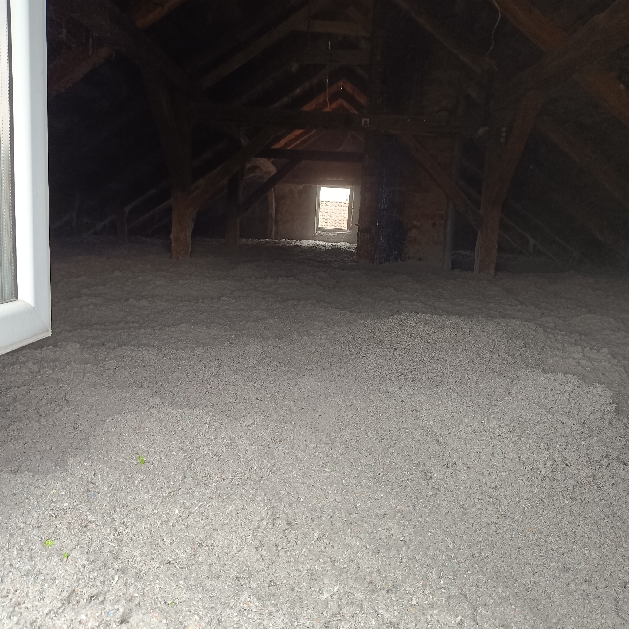 Poddasze wypełnione szarą, sypką izolacją, widoczne drewniane elementy konstrukcyjne dachu i okno w tle.