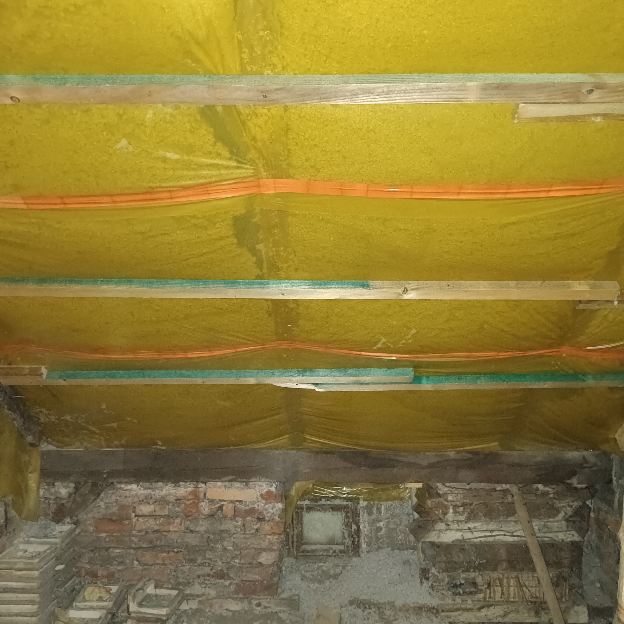 Żółta folia izolacyjna zamontowana pod drewnianymi belkami stropu poddasza, widoczne pomarańczowe kable i fragment murowanej ściany z cegieł.