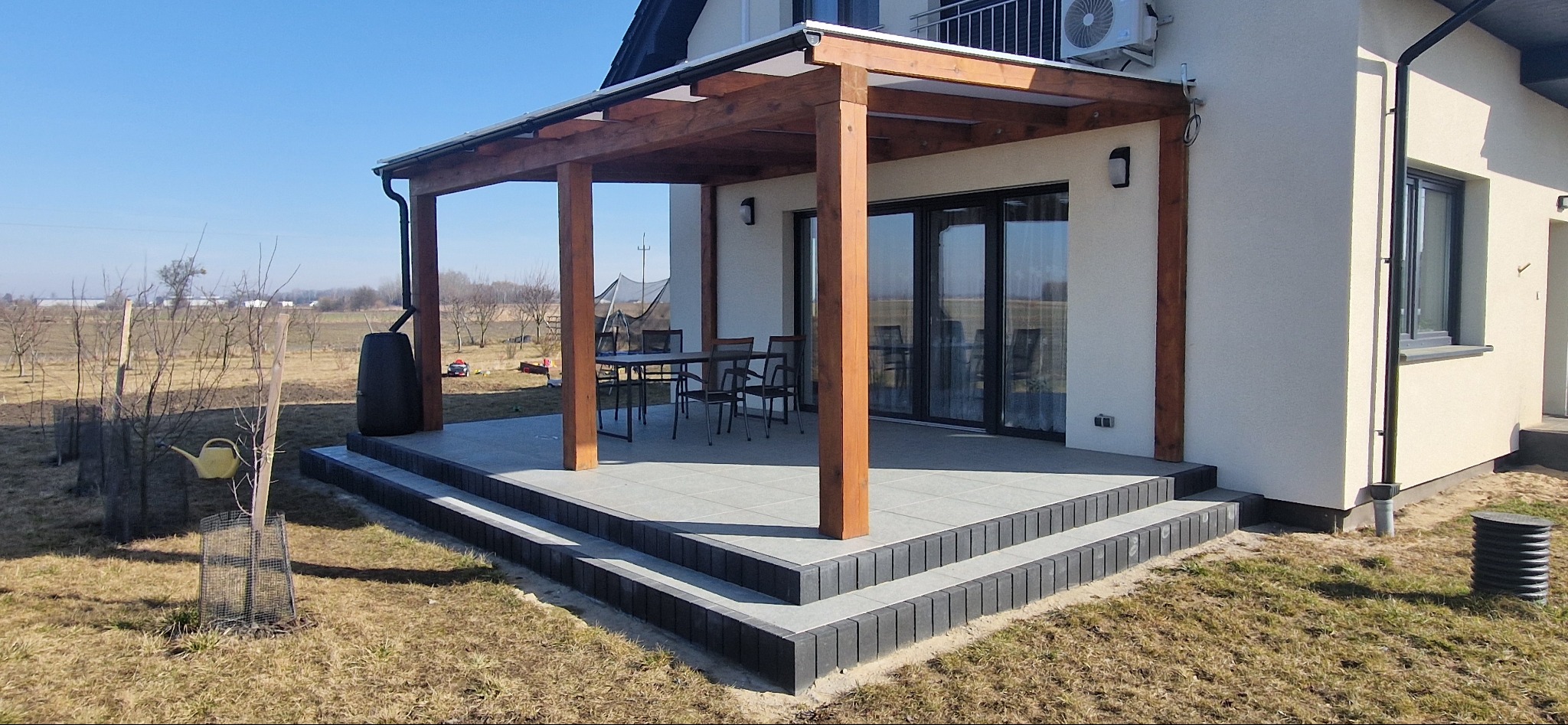 Drewniana pergola zadaszona, przylegająca do jasnego budynku z czarnymi oknami. Widoczne rynny, schody z kostki i elementy wyposażenia ogrodu.