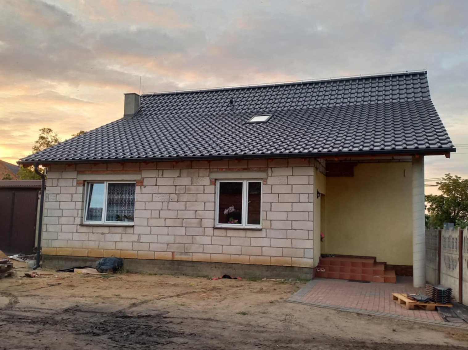 Dom jednorodzinny w trakcie budowy z ciemną dachówką, widoczne okna i fragment elewacji. Na pierwszym planie ziemia i materiały budowlane.