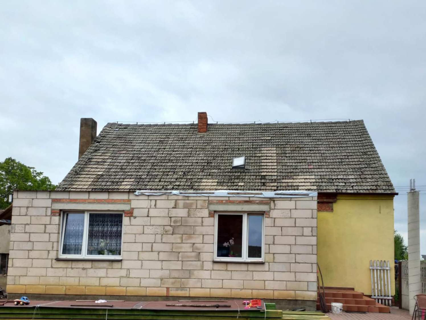Dom w trakcie budowy z widocznym dachem pokrytym starą dachówką. Część elewacji w surowym stanie, druga pomalowana na żółto. Widoczne materiały budowlane.
