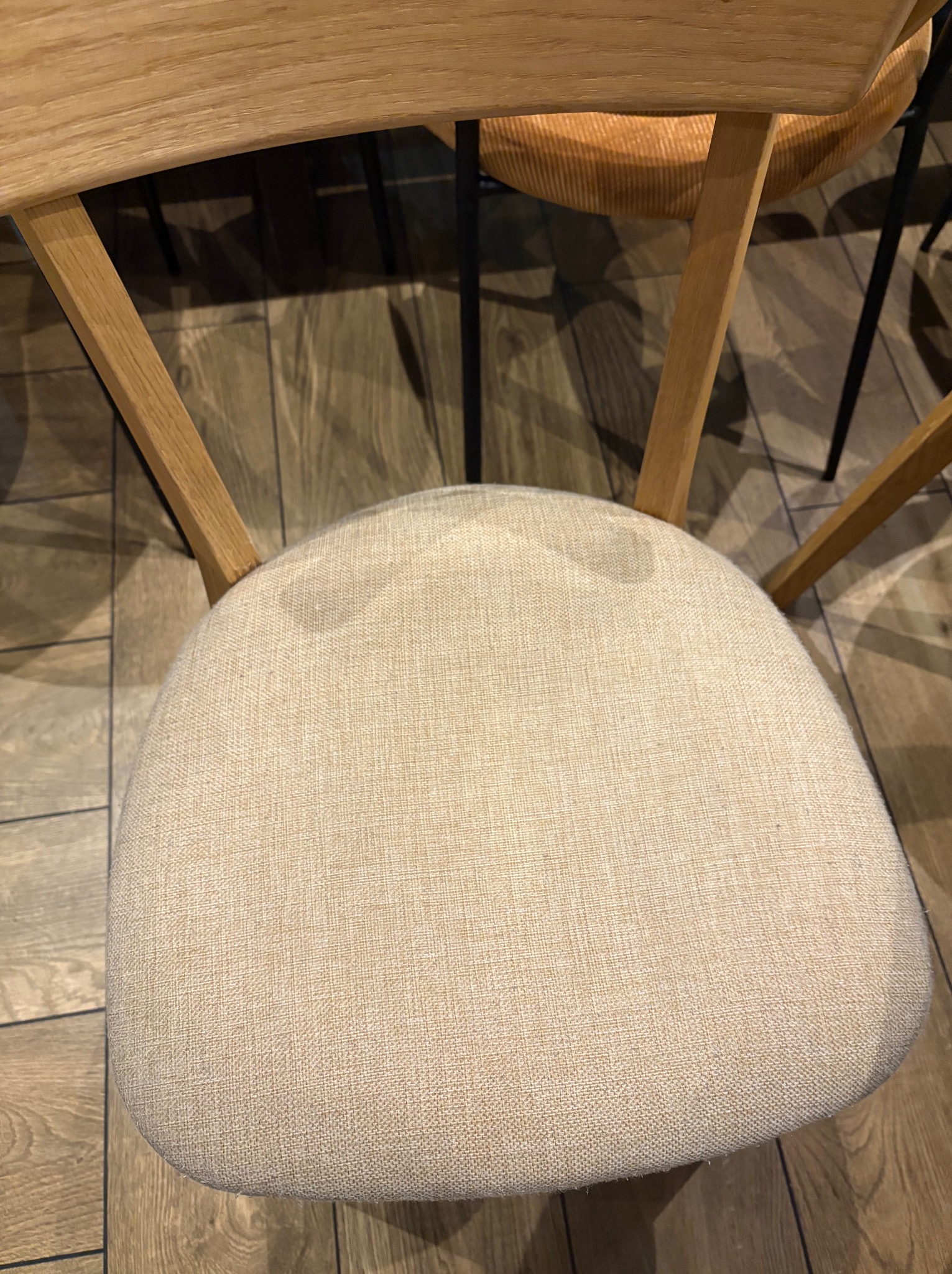 Krzesło z jasną tapicerką widziane z góry, z widocznymi plamami na siedzisku. Drewniana konstrukcja krzesła na tle podłogi z paneli.