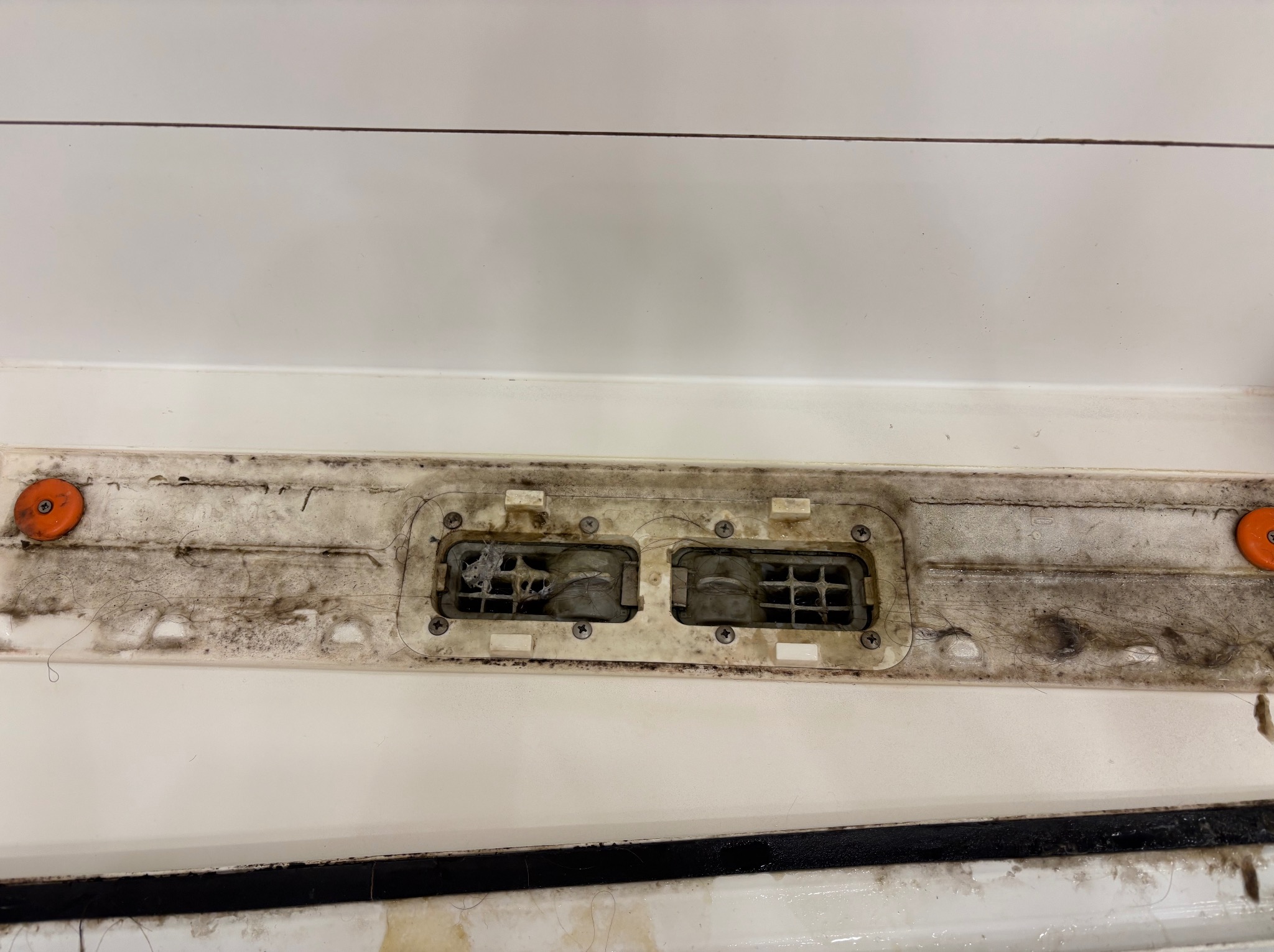 Zanieczyszczona kratka wentylacyjna w łazience, widoczne osady kurzu i włosów. Biała powierzchnia wokół kratki z plamami i zabrudzeniami. Dwa pomarańczowe elementy mocujące.