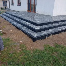 GRUPA AKORES SPÓŁKA Z OGRANICZONĄ ODPOWIEDZIALNOŚCIĄ - Nowo ułożone, szare, prostokątne płyty chodnikowe tworzą schody i taras przed domem z brązowymi drzwiami balkonowymi; widoczna świeżo zasiana trawa i fragment ubrania wykonawcy.