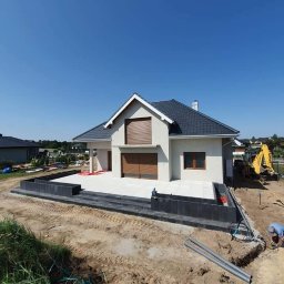 GRUPA AKORES SPÓŁKA Z OGRANICZONĄ ODPOWIEDZIALNOŚCIĄ - Nowoczesny dom jednorodzinny z wykończonym tarasem z jasnych płyt i obrzeżami z ciemnej kostki, widoczny w trakcie prac ziemnych na posesji, z koparką w tle.