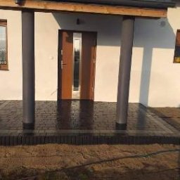 GRUPA AKORES SPÓŁKA Z OGRANICZONĄ ODPOWIEDZIALNOŚCIĄ - Nowo ułożona, ciemnobrązowa kostka brukowa przed wejściem do domu z dwoma kolumnami podtrzymującymi zadaszenie. Widoczne świeże fugi i minimalne zabrudzenia piaskiem.