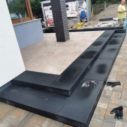 GRUPA AKORES SPÓŁKA Z OGRANICZONĄ ODPOWIEDZIALNOŚCIĄ - Czarne, gładkie stopnie z kostki brukowej prowadzące do domu, z widocznymi śladami po pracy i rękawicami roboczymi, w tle kontener budowlany i przygotowany teren pod dalsze prace.