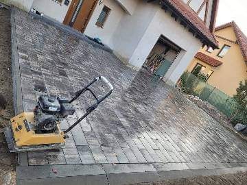 Świeżo ułożona, szara kostka brukowa na podjeździe przed domem, częściowo ubita zagęszczarką. Widoczny fragment elewacji domu z jasną fasadą i brązowymi drzwiami.