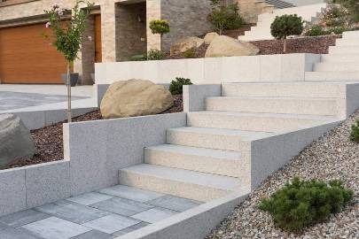 Schody zewnętrzne z jasnego kamienia prowadzące do domu, otoczone kamieniami i roślinami, z widoczną kostką brukową w dolnej części kadru.