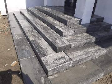 Schody zewnętrzne wyłożone szarą kostką brukową imitującą drewno, z widocznymi fugami i nierównościami terenu w tle.