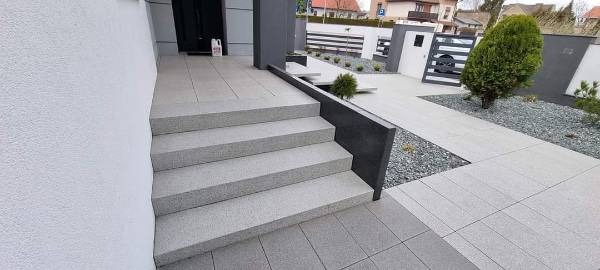 Nowoczesne schody wejściowe i chodnik wyłożone jasnoszarą kostką brukową, z elementami dekoracyjnymi z drobnego grysu, prowadzące do domu z białym tynkiem i antracytowymi detalami.