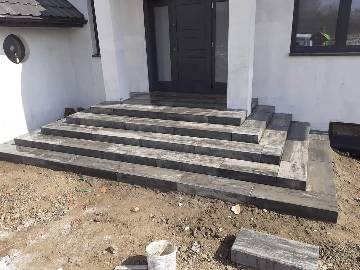 Schody wejściowe do domu wyłożone szarą kostką brukową, widoczne niedokończone prace i rozsypany piasek. Ujęcie z dołu, z widoczną elewacją budynku.