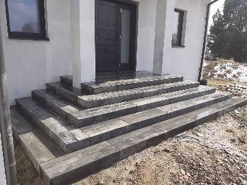 Schody wejściowe do domu wyłożone szarą kostką brukową, widoczne także fragment elewacji budynku, okno i drzwi wejściowe. Na ziemi resztki śniegu.