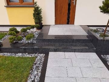Schody i ścieżka z jasnoszarej kostki granitowej prowadzące do drzwi wejściowych domu, obrzeża z ciemnego granitu, ozdobne nasadzenia iglaków i trawnika z kamiennym obrzeżem.