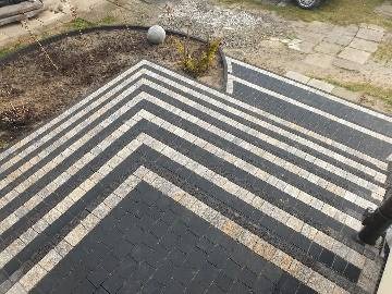 Schody zewnętrzne wyłożone kostką brukową w odcieniach szarości i beżu, z widoczną precyzją ułożenia wzoru. Na górze schodów widoczny fragment trawnika z elementami dekoracyjnymi, w tle fragment...