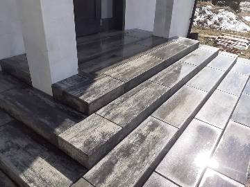 Schody wejściowe wyłożone szarą kostką brukową o nieregularnej fakturze, z widocznym odbiciem światła słonecznego na powierzchni kostki. Białe filary po bokach wejścia.