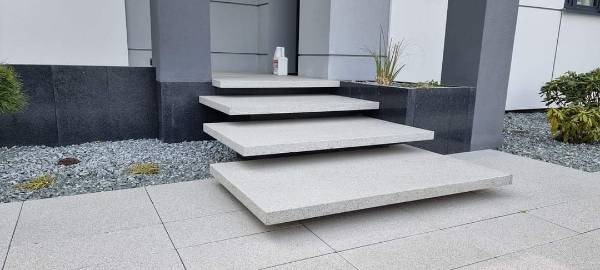 Nowoczesne, minimalistyczne schody zewnętrzne z jasnoszarego kamienia, prowadzące do wejścia budynku. Widoczne płyty chodnikowe w tym samym odcieniu, drobny żwir dekoracyjny i fragmenty roślinności.