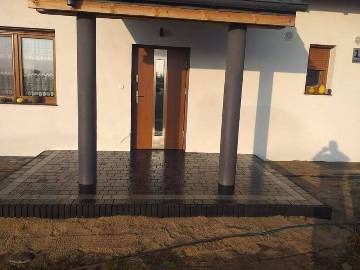 Nowo ułożona, ciemnobrązowa kostka brukowa przed wejściem do domu z dwoma kolumnami podtrzymującymi zadaszenie. Widoczne świeże fugi i minimalne zabrudzenia piaskiem.