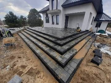 Ukończone schody wejściowe do domu wyłożone ciemnoszarą kostką brukową, widoczne narzędzia i resztki piasku, częściowo ułożona nawierzchnia przed domem, pochmurne niebo w tle.