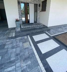 Nowoczesne podejście do domu wyłożone szarą kostką brukową z białymi płytami przejściowymi, donica z kwiatami przy wejściu, widok na drzwi wejściowe i fragment elewacji budynku.