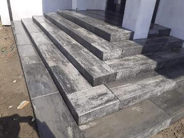 Schody zewnętrzne wyłożone ciemnoszarą kostką brukową o nieregularnej fakturze, prowadzące do budynku w Toruniu. Widoczne ślady świeżego cementu. Częściowo ukończona realizacja.