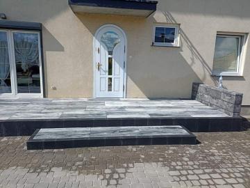 Wyłożony kostką brukową taras przed domem z białymi drzwiami wejściowymi. Kostka w odcieniach szarości i czerni, ułożona w geometryczny wzór. Widoczny fragment chodnika z kostki brukowej w tle.