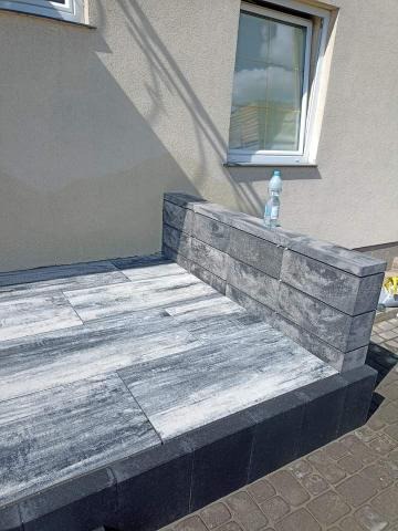 Wykończony taras z szarych płyt imitujących drewno, obłożony ciemną obwódką, z niskim murem z szarych bloczków betonowych. Na murku stoi butelka wody.