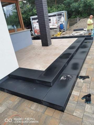 Czarne, gładkie stopnie z kostki brukowej prowadzące do domu, z widocznymi śladami po pracy i rękawicami roboczymi, w tle kontener budowlany i przygotowany teren pod dalsze prace.