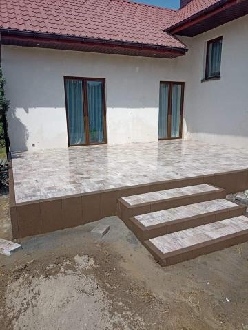 Wyłożony kostką brukową taras z trzema schodami prowadzącymi do domu z czerwonym dachem, widoczne niedokończone prace ziemne wokół.