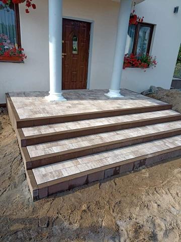 Schody wejściowe do domu wyłożone kostką brukową w odcieniach szarości i brązu, z kolumnami i kwiatami w oknach w tle.