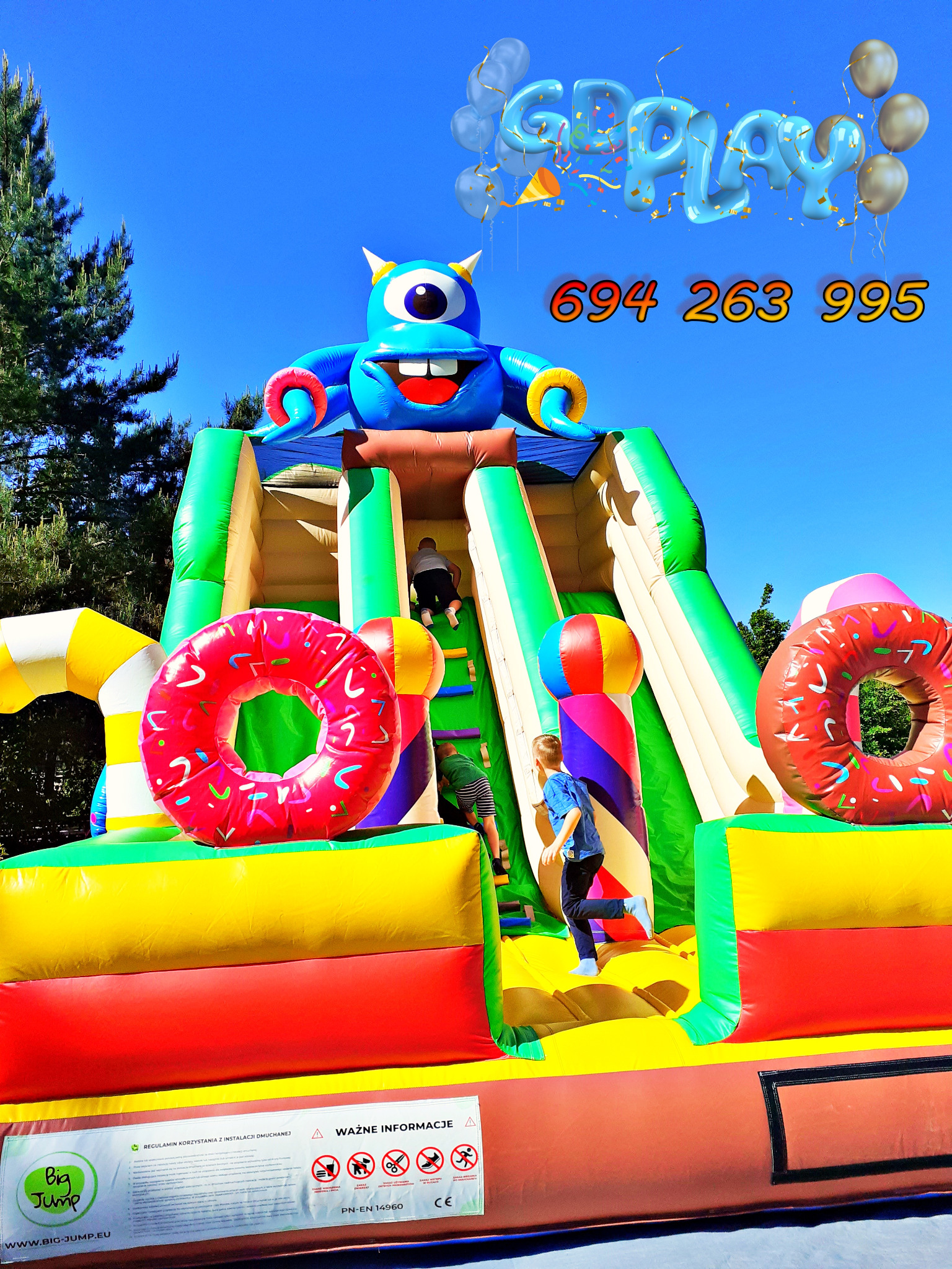 Kolorowa, dmuchana zjeżdżalnia w kształcie potwora z jednym okiem, z dwoma torami zjazdowymi, udekorowana dmuchanymi pączkami, dzieci wchodzą i zjeżdżają, na tle błękitnego nieba z napisem 'Goplay'...