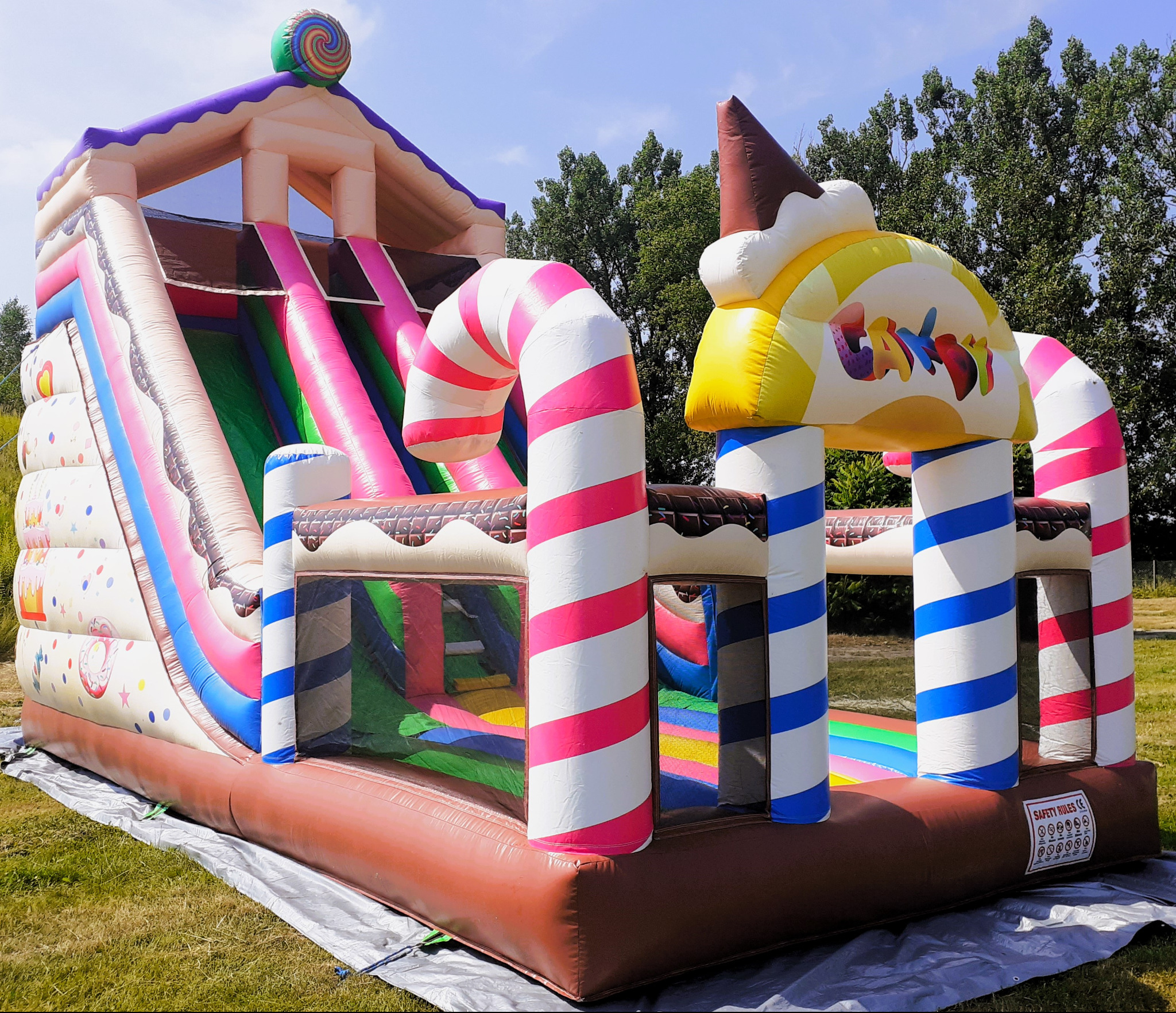 Kolorowa, dmuchana zjeżdżalnia w kształcie słodyczy, z wysoką ślizgawką, elementami przypominającymi lizaki i lodowe rożki, ustawiona na trawie w słoneczny dzień.