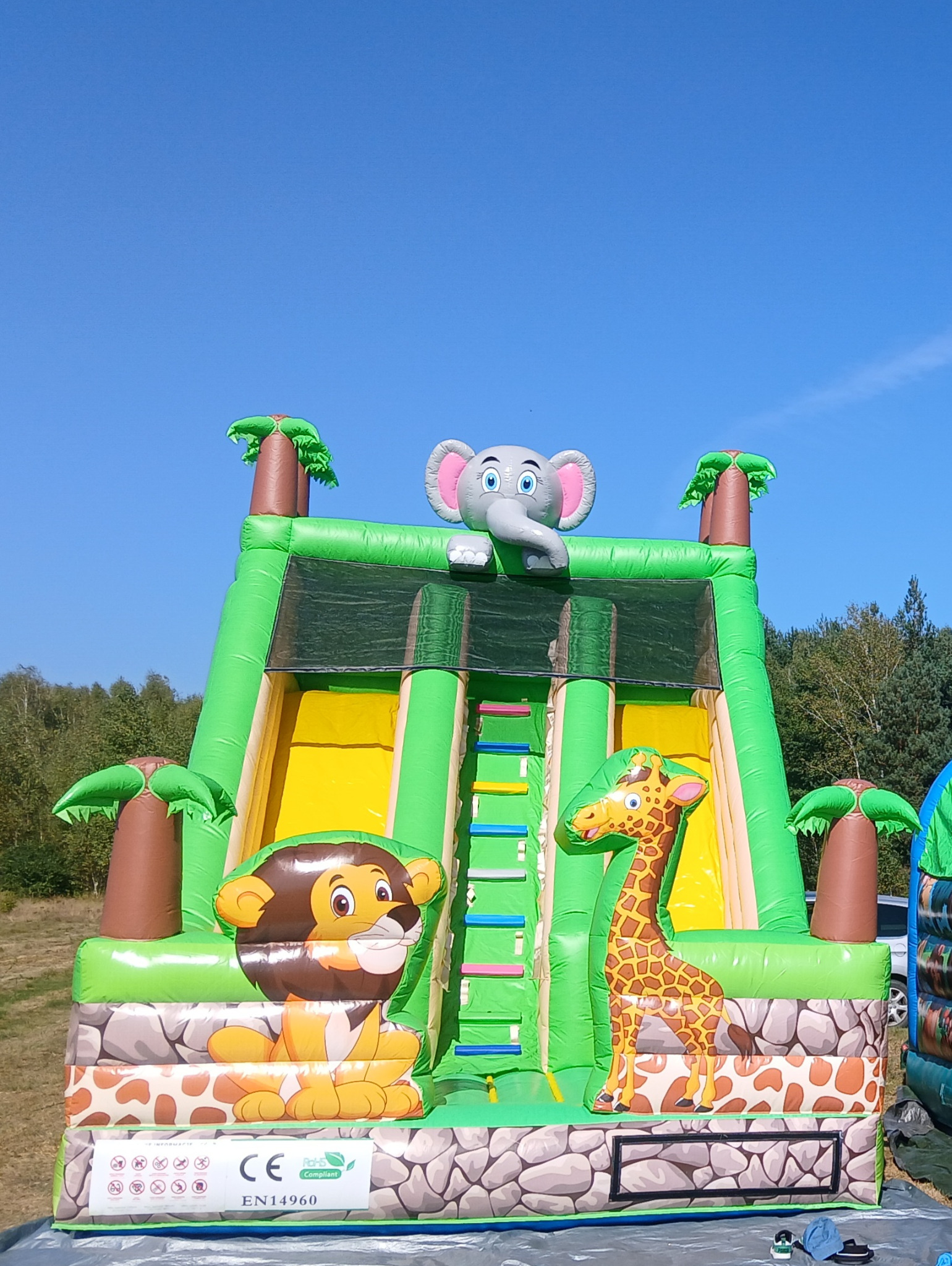 Nadmuchiwana zjeżdżalnia w kształcie dżungli z motywami zwierzęcymi, takimi jak słoń na szczycie, lew i żyrafa po bokach, ustawiona na trawiastym terenie pod błękitnym niebem.