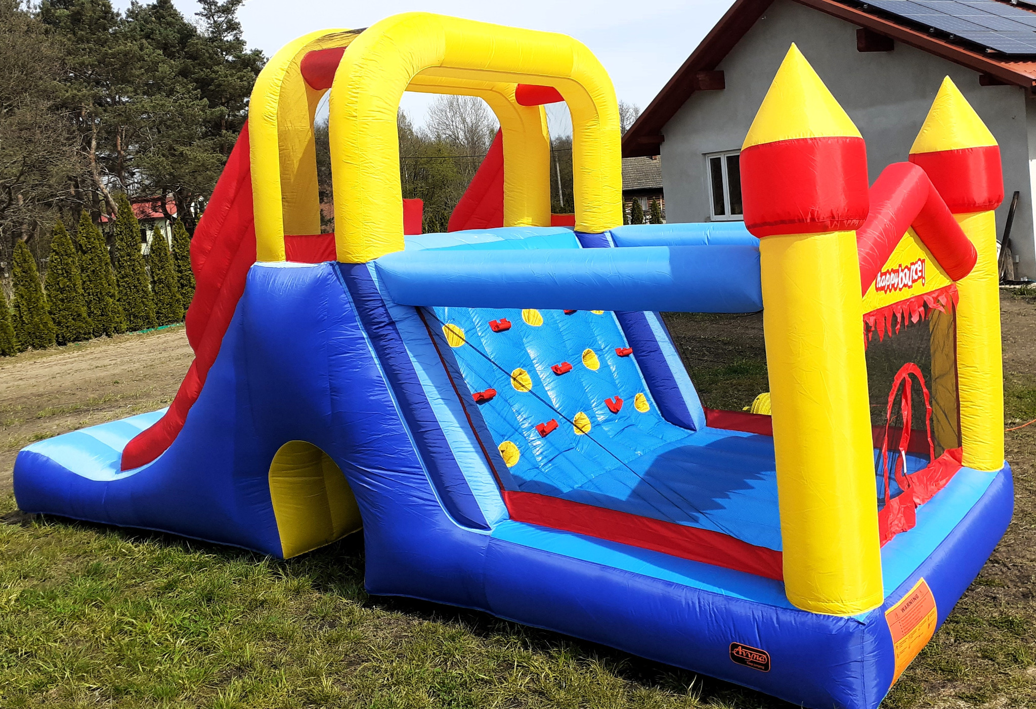 Kolorowy, dmuchany plac zabaw dla dzieci z elementami zjeżdżalni, ścianki wspinaczkowej i małego zamku, ustawiony na trawie przed domem.