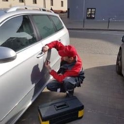 Alex Kowal - Mężczyzna w czerwonej czapce i kombinezonie naprawia klamkę w srebrnym samochodzie kombi na ulicy w Brwinowie, obok stoi czarna skrzynka z narzędziami.