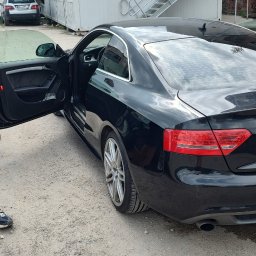 Alex Kowal - Czarne Audi A5 z otwartymi drzwiami kierowcy, zaparkowane na żwirowym podłożu przed budynkiem kontenerowym z metalowymi schodami.