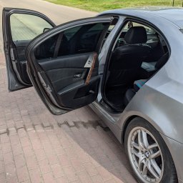 Alex Kowal - Srebrny samochód z otwartymi drzwiami na brukowanym chodniku, widoczny fragment wnętrza z czarną tapicerką i drewnianym wykończeniem. W tle budynki mieszkalne i drzewa.