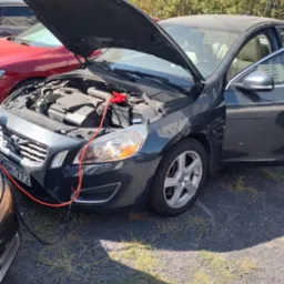 Szary samochód Volvo z otwartą maską, podłączony kablem rozruchowym do akumulatora innego pojazdu, stojący na żwirowym parkingu w słoneczny dzień.