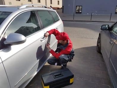 Mężczyzna w czerwonej czapce i kombinezonie naprawia klamkę w srebrnym samochodzie kombi na ulicy w Brwinowie, obok stoi czarna skrzynka z narzędziami.