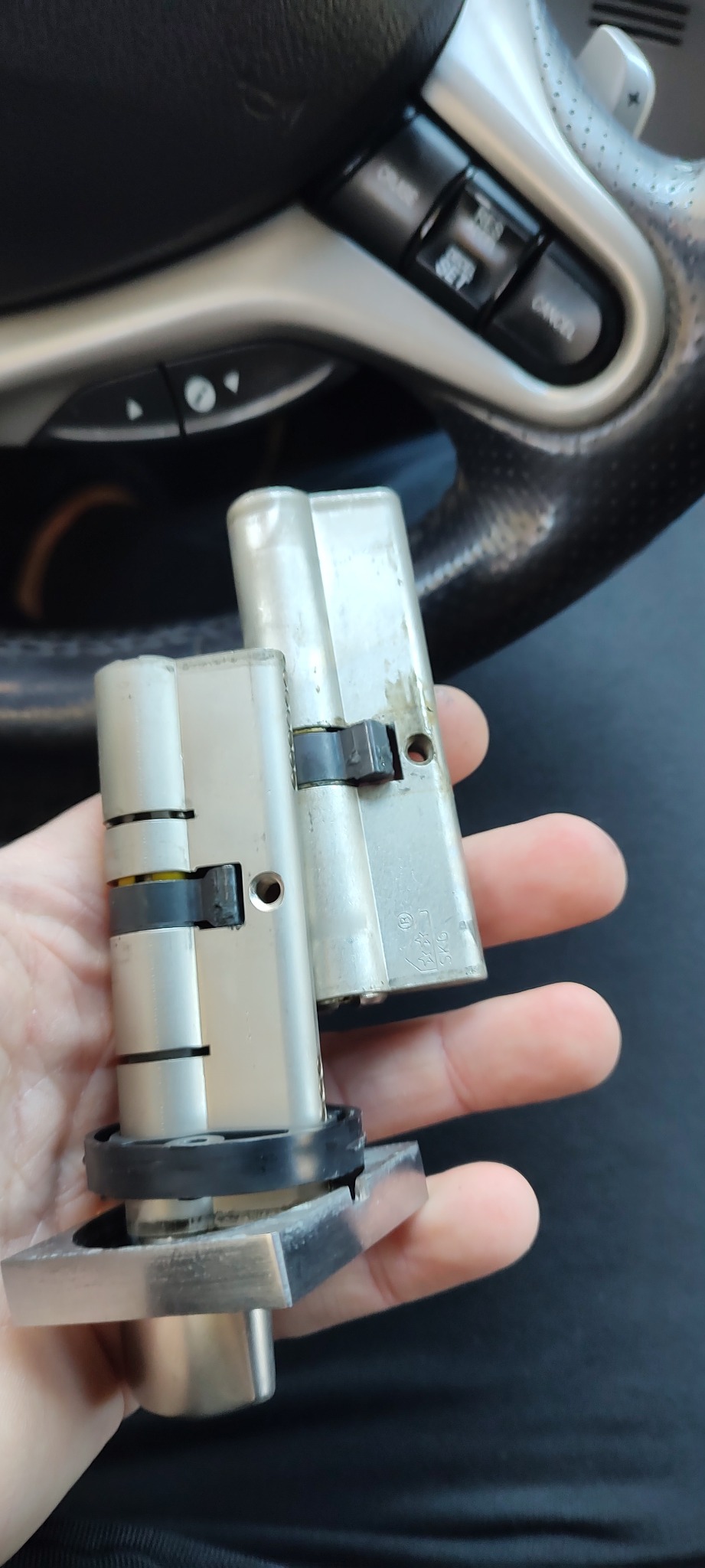 Dwa używane, srebrne zamki bębenkowe do drzwi, trzymane w dłoni na tle kierownicy samochodowej.