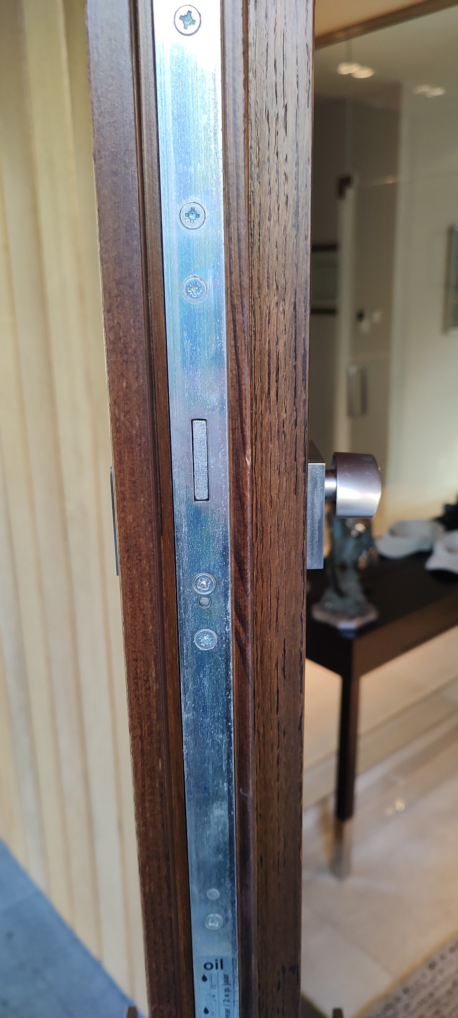 Szczegółowe ujęcie zamka w drewnianych drzwiach, widoczna srebrna listwa z wkrętami i instrukcją smarowania, z brązową gałką zamka po prawej stronie, na tle rozmytego wnętrza.