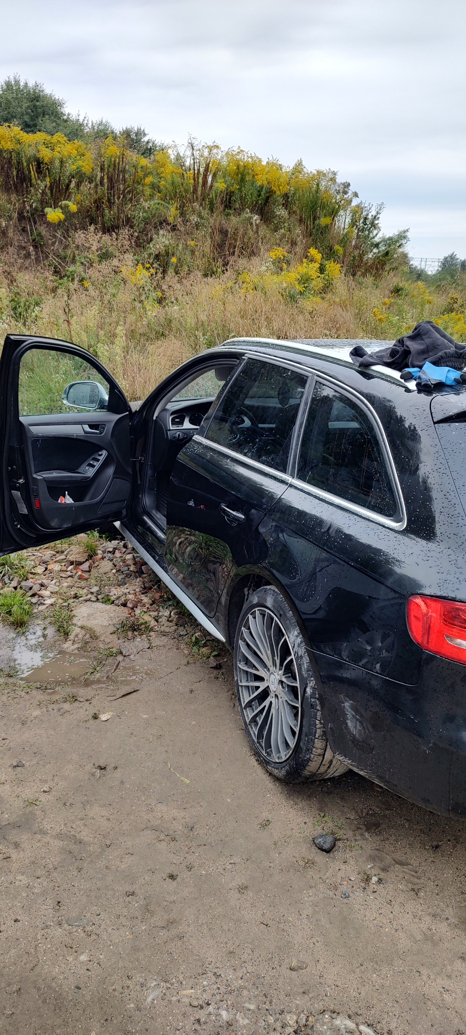 Czarny samochód kombi z otwartymi drzwiami stojący na błotnistej drodze polnej, z widocznymi śladami błota na karoserii i oponach. Na dachu leżą ciemne ubrania. W tle widoczna roślinność.